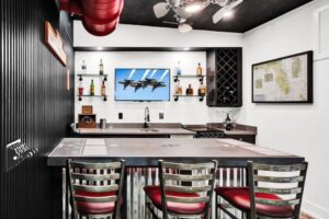 Aviation themed kitchen and dining area inside a hangar home built by Newberry, designed to also accommodate small aircraft. A bar-height counter with a runway-marked concrete top faces four metal barstools with red cushioned seats. The counter front is clad in corrugated metal panels. Behind the bar, open shelving displays liquor bottles, a wine rack cabinet holds bottles at an angle, and a wall-mounted TV shows the Blue Angels in formation flight. A utility sink with a gooseneck faucet is set into the dark countertop. An aeronautical map of Florida is framed on the right wall. A ceiling fan with a light kit is mounted on the black-painted ceiling, and a partial view of a red aircraft propeller or nose is visible in the upper left corner.