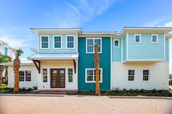 Exterior front view of The Duval, a two-story coastal-style home in the Ponce Inlet Key development in Ponce Inlet, Florida. The home features light blue and teal lap siding accents on a white stucco base. The entrance features a covered porch with a metal roof, wood bracket details, and glass-paned double doors. Two tall palm trees flank the entryway, and fresh landscaping lines the foundation. Multiple white-trimmed windows are visible across both floors. A brick paver driveway leads up to the home under a partly cloudy blue sky.
