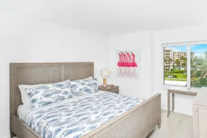 After bedroom remodel with bright finishes, updated flooring, and coastal-inspired design by Newberry Homes Large windows facing large grass area. Flamingo painting on wall next to window.
