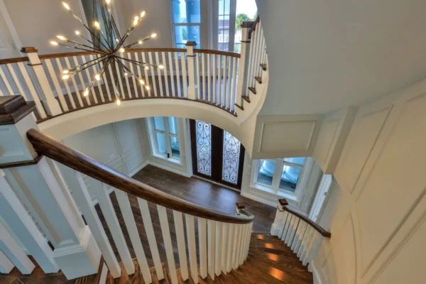 A high-angle view from an upper landing looking down into a grand entryway featuring a sweeping curved staircase with dark wood handrails and white spindles. A large, modern sputnik-style chandelier hangs over the space, illuminating dark hardwood floors and a set of double entry doors with decorative iron scrollwork.