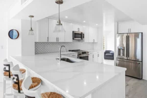 Open concept condo kitchen after Newberry Homes renovation with improved flow and modern materials in New Smyrna Beach, Florida. Wicker bar stools up against kitchen counter, view of stainless steal french door refrigerator, stainless steels stove and oven, stainless steel microwave in all white cabinetry.
