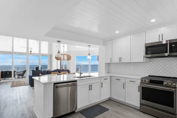 Coastal-inspired condo kitchen renovation by Newberry Homes with clean lines and modern upgrades in New Smyrna Beach, Florida. View of kitchen - looking at kitchen bar with two hanging light fixtures, stainless steel dishwasher, white cabinetry, stainless steel microwave, stainless steel stove and oven, looking at living room with large floor to ceiling windows with a large overzied balcony and a ocean view.