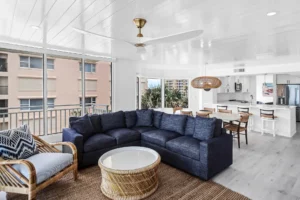 After photo of renovated condo kitchen and dining area with cohesive design and improved functionality in New Smyrna Beach, Florida by Newberry homes. Large oversized L shape couch, wicker table with oversized wicker chair and cushion. Beyond the couch is a large kitchen table facing three windows with a wicker light fixture. The open kitchen has a bar, and stainless steel refrigerator with french doors.
