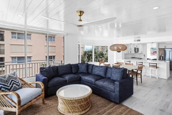 After photo of renovated condo kitchen and dining area with cohesive design and improved functionality in New Smyrna Beach, Florida by Newberry homes. Large oversized L shape couch, wicker table with oversized wicker chair and cushion. Beyond the couch is a large kitchen table facing three windows with a wicker light fixture. The open kitchen has a bar, and stainless steel refrigerator with french doors.
