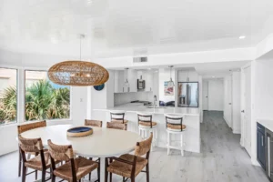 After photo of modern condo kitchen renovation by Newberry Homes with white cabinetry, updated countertops, with large oversized windows in front of the kitchen table with wicker style chairs and wicker light fixture, and wicker style chairs for kitchen bar. A new and improved design layout in New Smyrna Beach, Florida.
