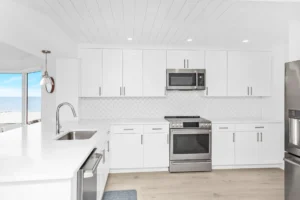 Open concept condo kitchen after renovation by Newberry Homes with improved flow and modern materials in New Smyrna Beach, Florida. View of stainless steel dishwasher, microwave, stove, oven and refrigerator. Large oversized windows