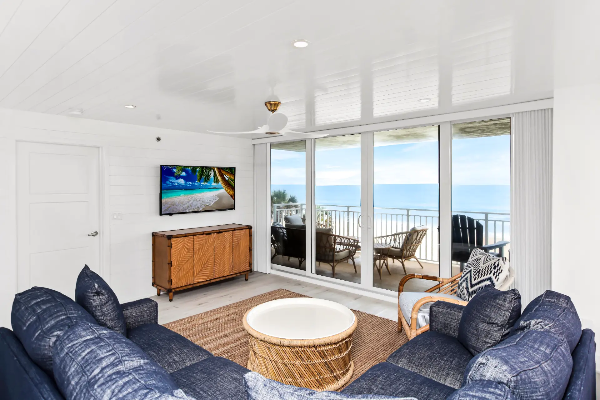 Oceanfront condo living room renovation after completion in New Smyrna Beach, Florida, featuring coastal finishes, white shiplap walls, and panoramic ocean views by Newberry Homes. TV cabinet with wooden accents, and a wicker style coffee table surrounded by an L shape blue couch.
