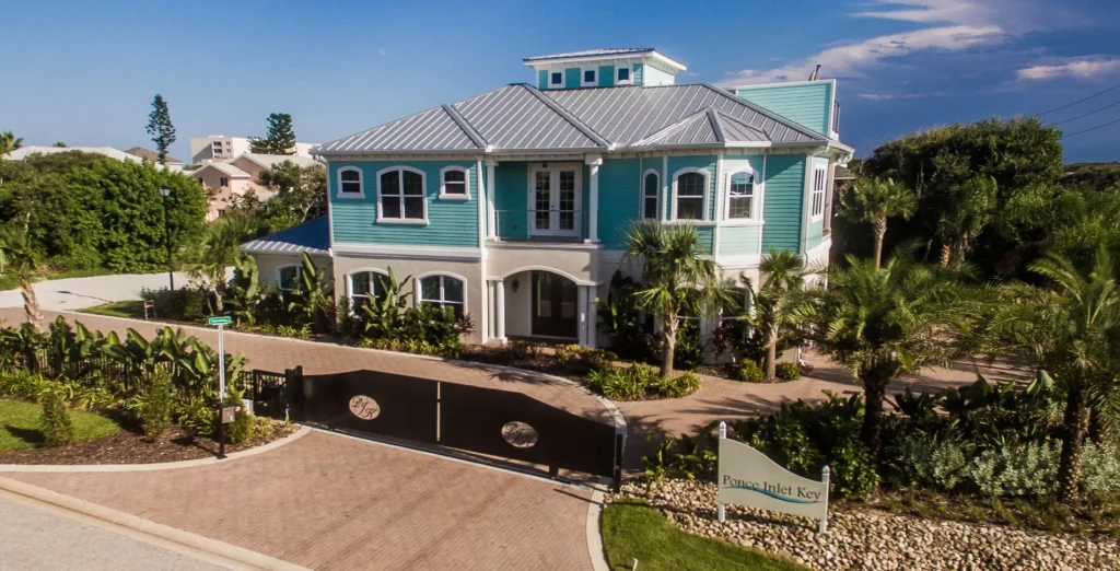 View of a coastal custom home with metal roof, tropical landscaping, and circular driveway in Ponce Inlet Florida By Newberry Homes. Ponce Inlet Key sign with metal gate surrounded by tropical landscaping around home.