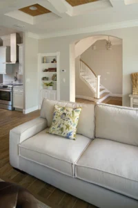 Living room with upholstered sofa, decorative pillows, wood-look flooring, and a custom arched opening connecting to the kitchen in a Florida design build home by Newberry Homes. Located in the Deltona–Daytona Beach–Ormond Beach Metropolitan Area
