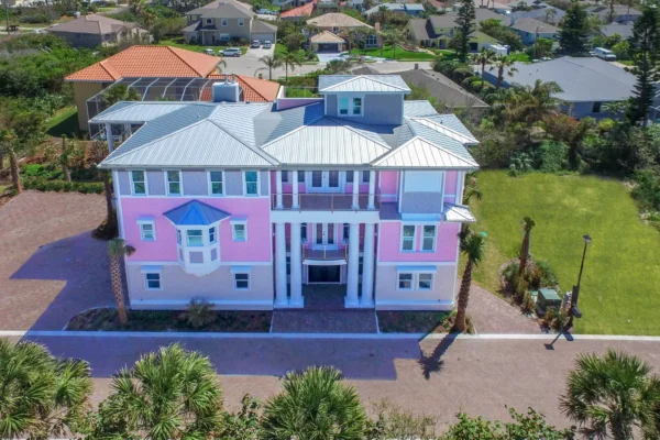 Front elevation of a custom coastal home in Deltona–Daytona Beach–Ormond Beach Metropolitan Area, with pastel exterior, metal roof, covered balconies, and brick paver driveway surrounded by tropical landscaping in Ponce Inlet Florida.