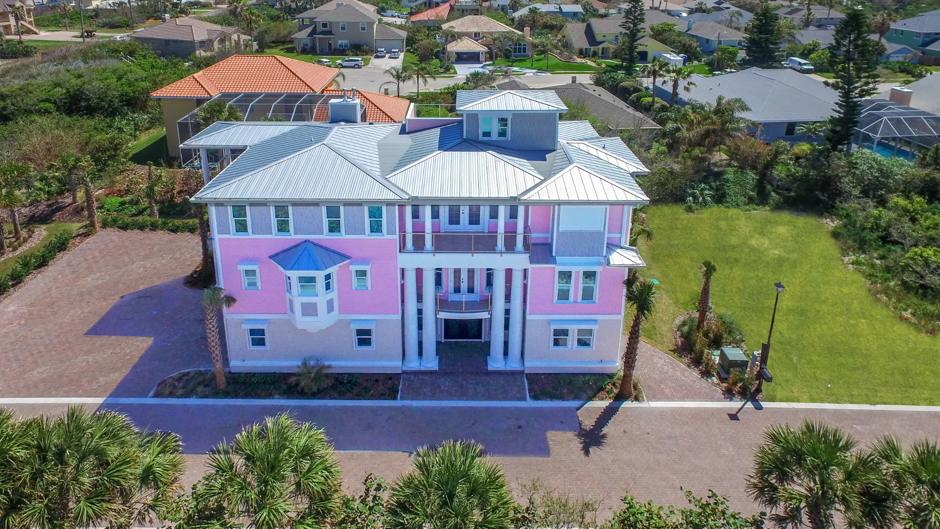 Front elevation of a custom coastal home in Deltona–Daytona Beach–Ormond Beach Metropolitan Area, with pastel exterior, metal roof, covered balconies, and brick paver driveway surrounded by tropical landscaping in Ponce Inlet Florida.