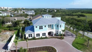 Front exterior of a gated coastal home with metal roof, pastel siding, and lush landscaping in Ponce Inlet Florida by Newberry Homes.