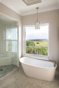 Freestanding soaking tub positioned beneath a large window with marsh views, glass-enclosed walk-in shower, marble tile flooring, and brass fixtures in a custom master bathroom in Ponce Inlet, Florida - the Deltona–Daytona Beach–Ormond Beach Metropolitan Area.