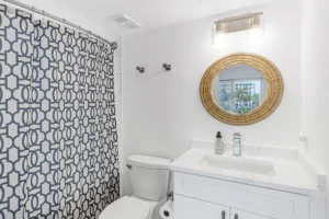 Remodeled guest bathroom with a white shaker vanity, quartz countertop, woven gold mirror, and navy patterned shower curtain in a Ponce Inlet condo by Newberry Homes.