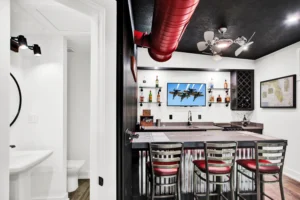 Aviation-themed wet bar in a Newberry Homes hangar home in Port Orange, Florida, with a runway-striped bar top, red metal barstools, corrugated steel paneling, red overhead ductwork, and a wall-mounted TV displaying the Blue Angels