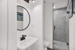 Bathroom in a Newberry Homes hangar home in Port Orange, Florida, featuring a white pedestal sink with matte black fixtures, round black-framed mirror, airplane artwork, and a glass walk-in shower with gray mosaic tile