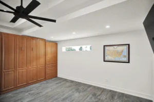 Bedroom in a Newberry Homes hangar home in Port Orange, Florida, featuring a built-in wood Murphy bed cabinet system, black ceiling fan, wood-look tile flooring, and a framed United States travel map on the wall
