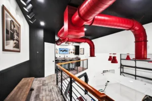 Mezzanine loft bar in a Newberry Homes hangar home in Port Orange, Florida, with bold red industrial ductwork, black shiplap accent wall, wood-look tile flooring, and a view down to an air hockey table