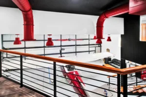 View from the mezzanine loft of a Newberry Homes hangar home in Port Orange, Florida, overlooking a red and white airplane parked beside a leather lounge seating area, with red ductwork and pendant lighting against black ceiling beams