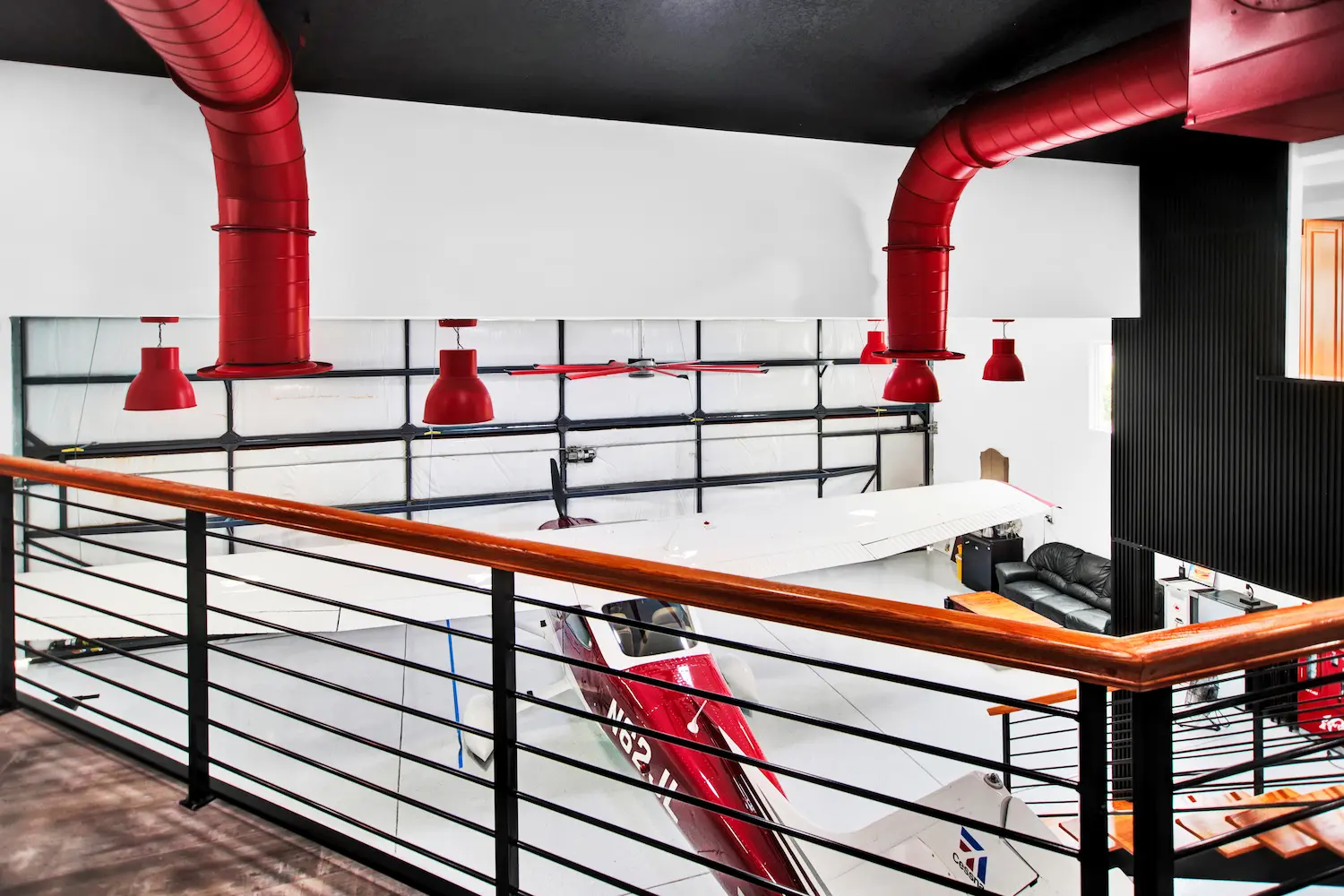 View from the mezzanine loft of a Newberry Homes hangar home in Port Orange, Florida, overlooking a red and white airplane parked beside a leather lounge seating area, with red ductwork and pendant lighting against black ceiling beams
