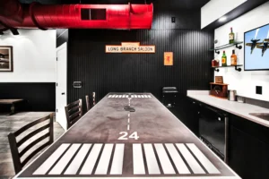Runway-themed dining table with painted taxiway markings and numbers in a Newberry Homes hangar home in Port Orange, Florida, beneath red industrial ductwork, with a black shiplap accent wall, Long Branch Saloon sign, and mini-bar in the background