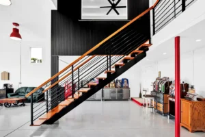 Wood and black steel staircase leading to a mezzanine loft in a Newberry Homes hangar home in Port Orange, Florida, with a vintage arcade and slot machine game room, leather sofa, and red pendant lighting below