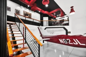 Two-story interior of a Newberry Homes hangar home in Port Orange, Florida, showing a wood and black steel staircase, mezzanine bar loft with red industrial ductwork, and the tail of a red and white Cessna Skylane parked below
