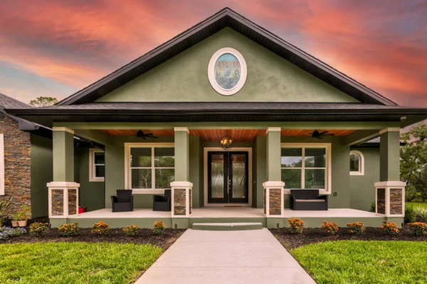 Luxury custom home exterior in Central Florida featuring a covered front porch, tapered columns with stone accents, green stucco facade, wood soffit ceiling, and landscaped entry at sunset.