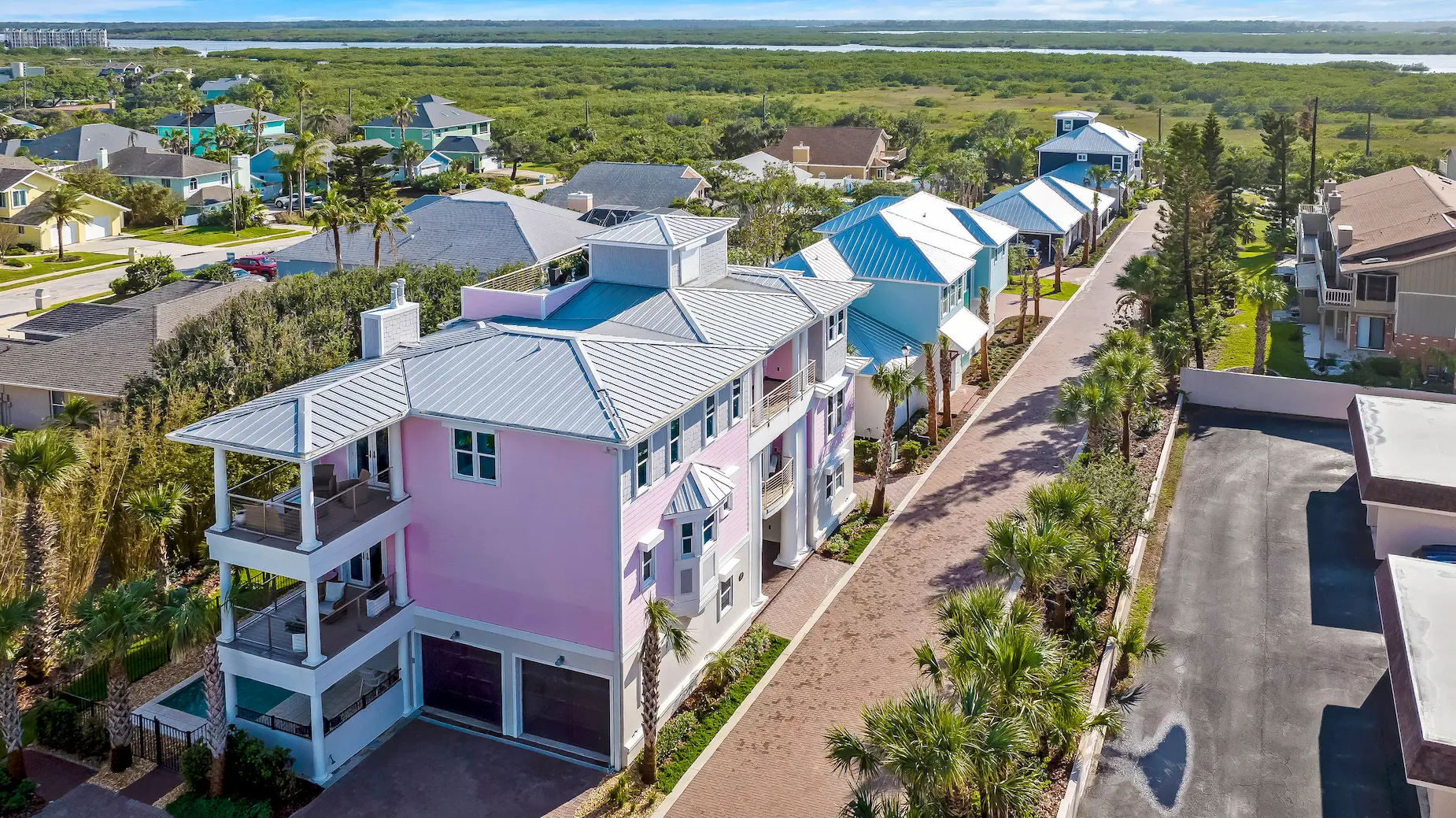Aerial view of a luxury custom home community in Ponce Inlet, Florida showing pastel-colored coastal homes with metal roofs, palm-lined private street, garages, and surrounding marshland in the background.