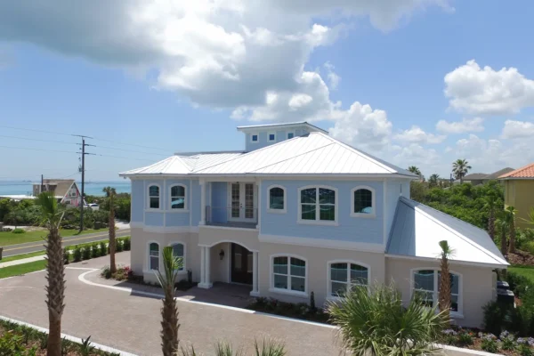 Luxury custom home exterior in Ponce Inlet, Florida featuring a light blue coastal-style residence with a white metal roof, arched windows, covered balcony, and paved driveway, viewed from the front at street level.
