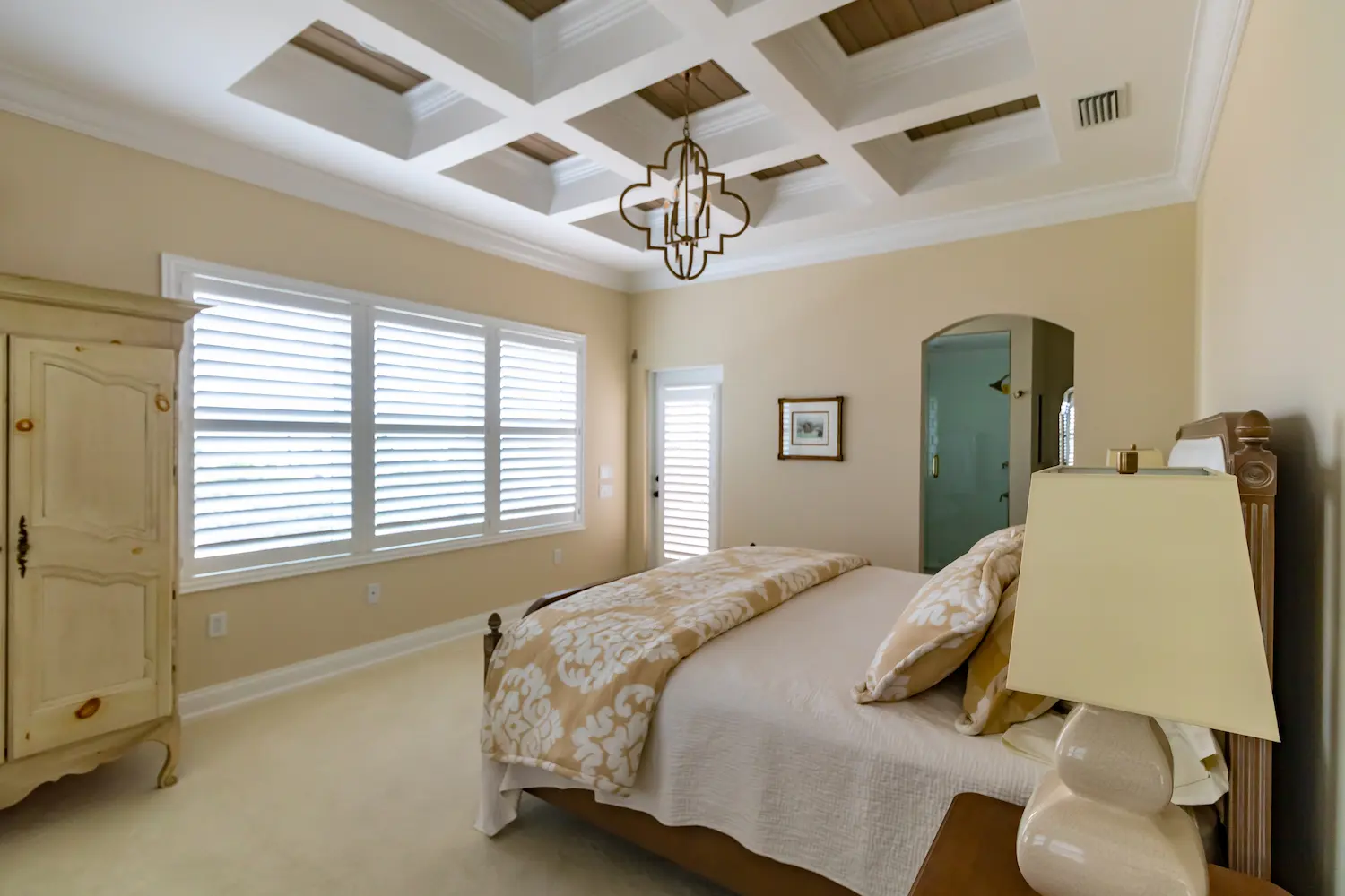 Luxury interior remodel bedroom in Ponce Inlet, Florida featuring a coffered ceiling, neutral color palette, plantation shutters, and custom trim details. Arch doorway