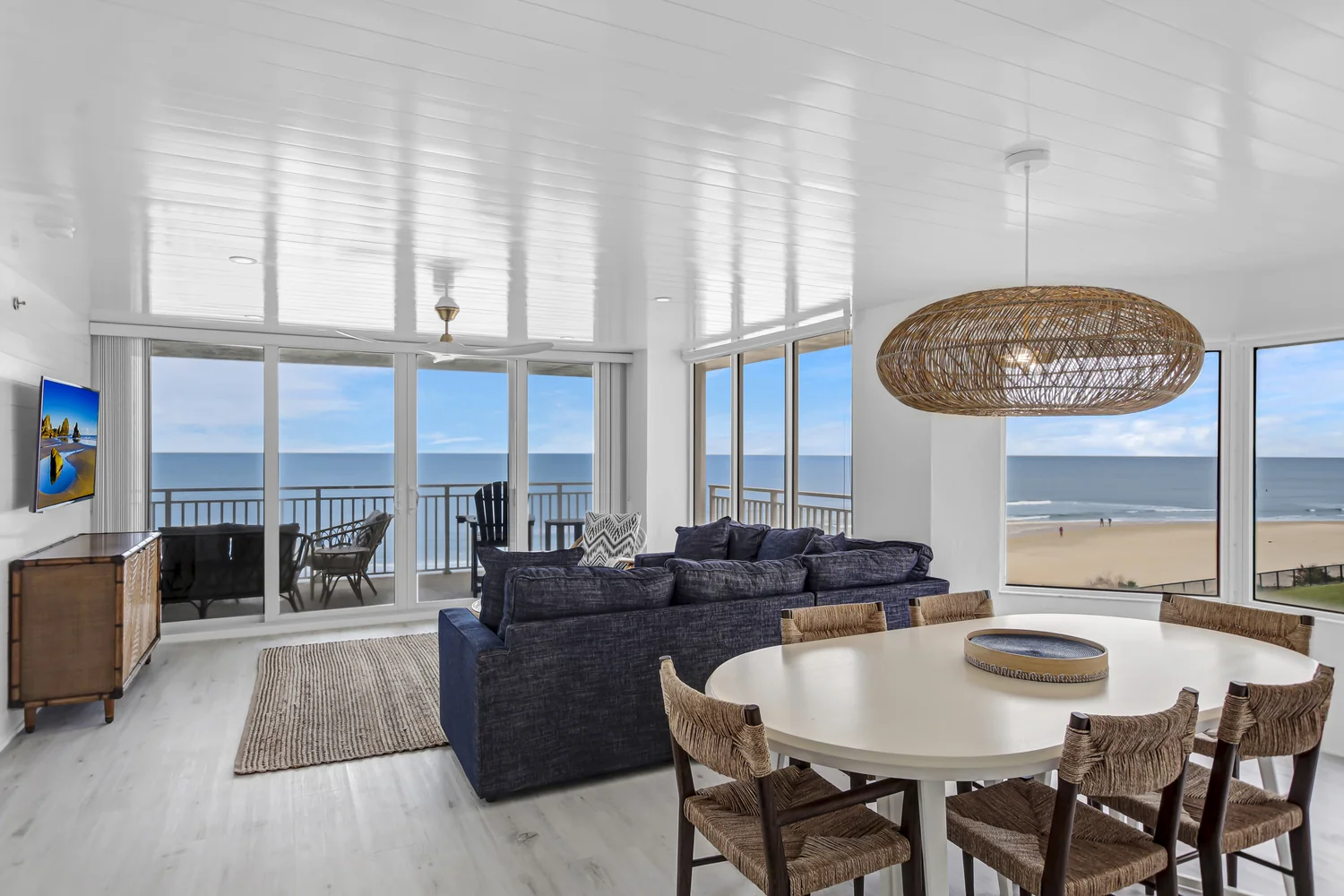 Oceanfront great room combining living and dining areas with wraparound ocean views, a navy sofa, and an oversized woven pendant light in a condo remodel by Newberry Homes in Ponce Inlet, Florida.