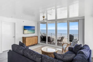 Oceanfront living room with a navy sectional, rattan accents, and sliding glass doors opening to a balcony with Atlantic Ocean views in a condo remodel by Newberry Homes in Ponce Inlet, Florida