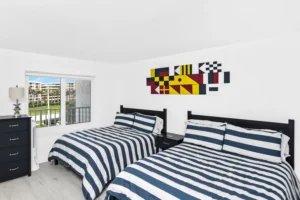 Guest bedroom with two navy and white striped beds, a black nightstand and dresser, and colorful nautical signal-flag wall art in a condo remodel by Newberry Homes in Ponce Inlet, Florida.