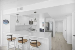 Bright white coastal kitchen with a quartz peninsula, counter stools, a scalloped tile backsplash, and stainless steel appliances in a condo remodel by Newberry Homes in Ponce Inlet, Florida.