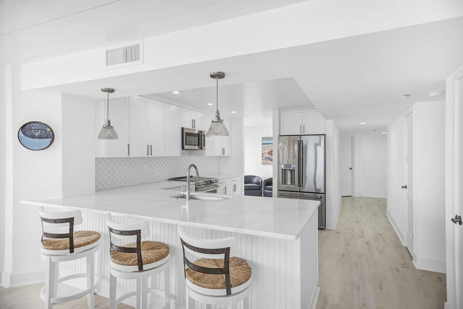 Bright white coastal kitchen with a quartz peninsula, counter stools, a scalloped tile backsplash, and stainless steel appliances in a condo remodel by Newberry Homes in Ponce Inlet, Florida.