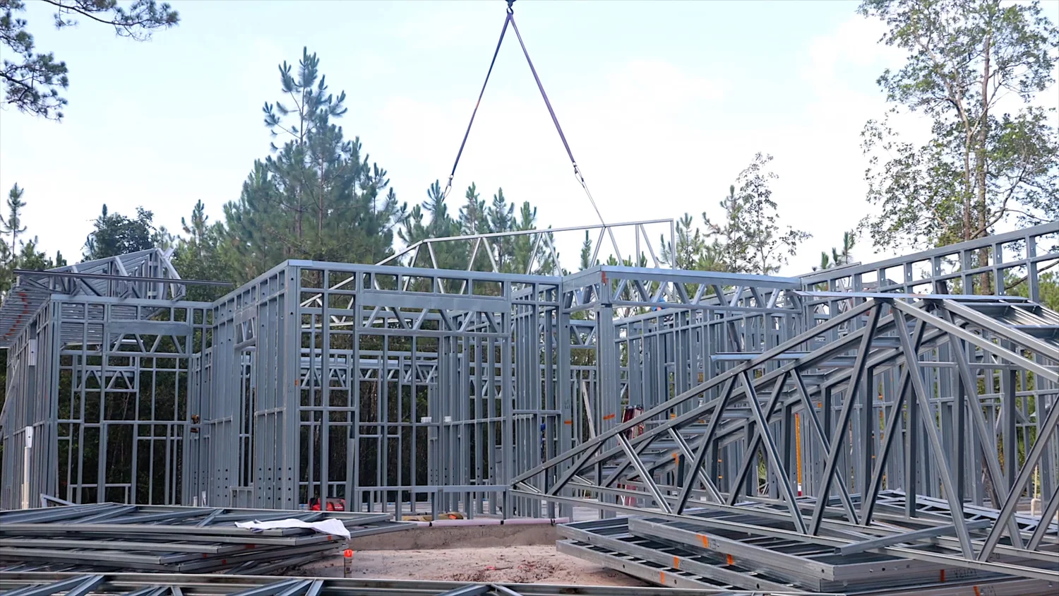 Crane lifting a steel roof truss into position over completed light gauge steel frame walls at a residential construction site surrounded by pine trees