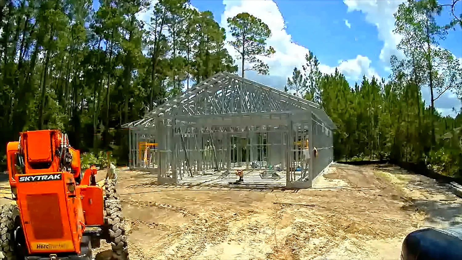 Completed light gauge steel frame residential home with full roof truss system installed, SkyTrak telehandler on site, surrounded by tall pine trees