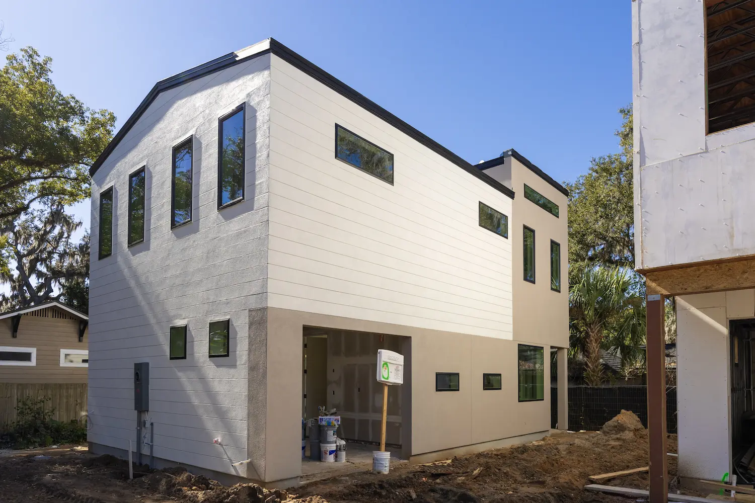 Rear construction progress view of a completed modern two-story home on W. Princeton Street in College Park, Orlando, featuring white horizontal fiber cement lap siding on the upper volume with black-framed windows, smooth gray stucco on the lower level, an open garage bay, paint supplies and construction materials at grade, and a partially clad adjacent unit visible to the right under mature oak trees